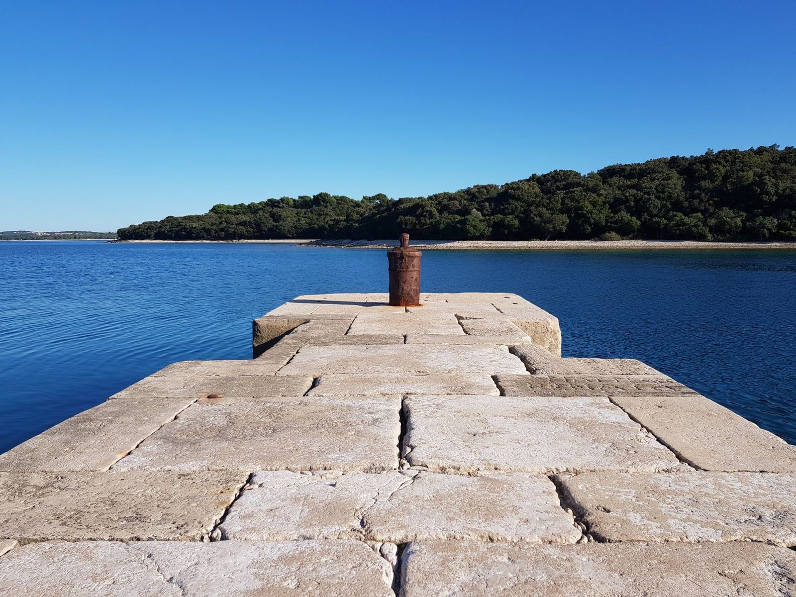 A stone pier with a weathered iron bollard extends into the sea in Fažana, overlooking the wooded islands of Brijuni National Park.